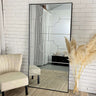 A large rectangular mirror with a black frame, leaning against a white brick wall, with a chair and decorative plant to its left.